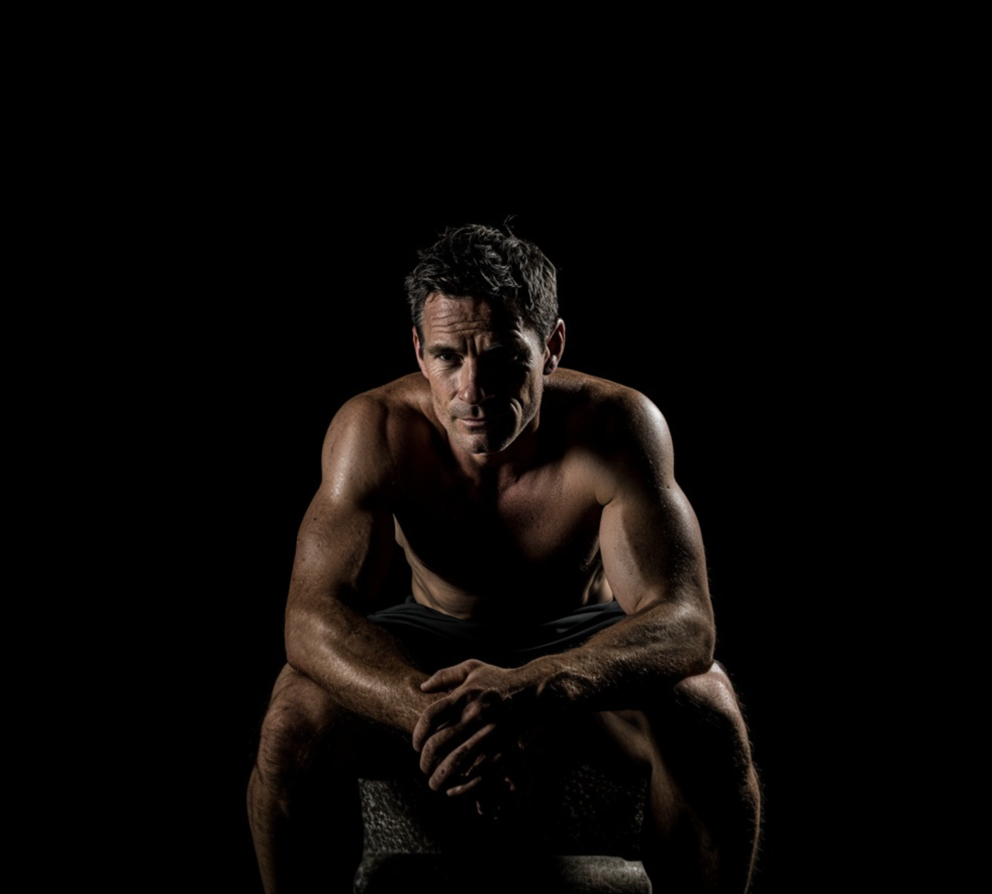 Shirtless man in his 30s sitting on a stone, looking directly at the camera against a dark background.